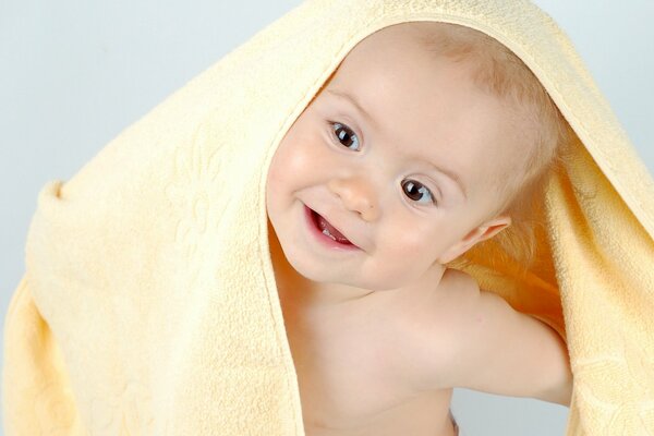 A baby peeking out from under a yellow terry towel