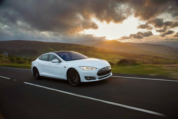Biała Tesla Model s na tle drogi, wzgórz i chmur