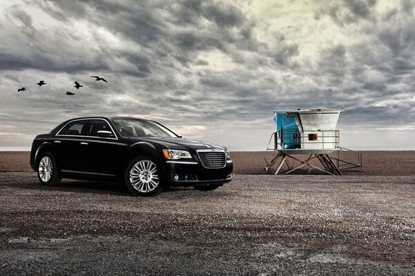 A black car is standing under clouds and birds are flying past this car