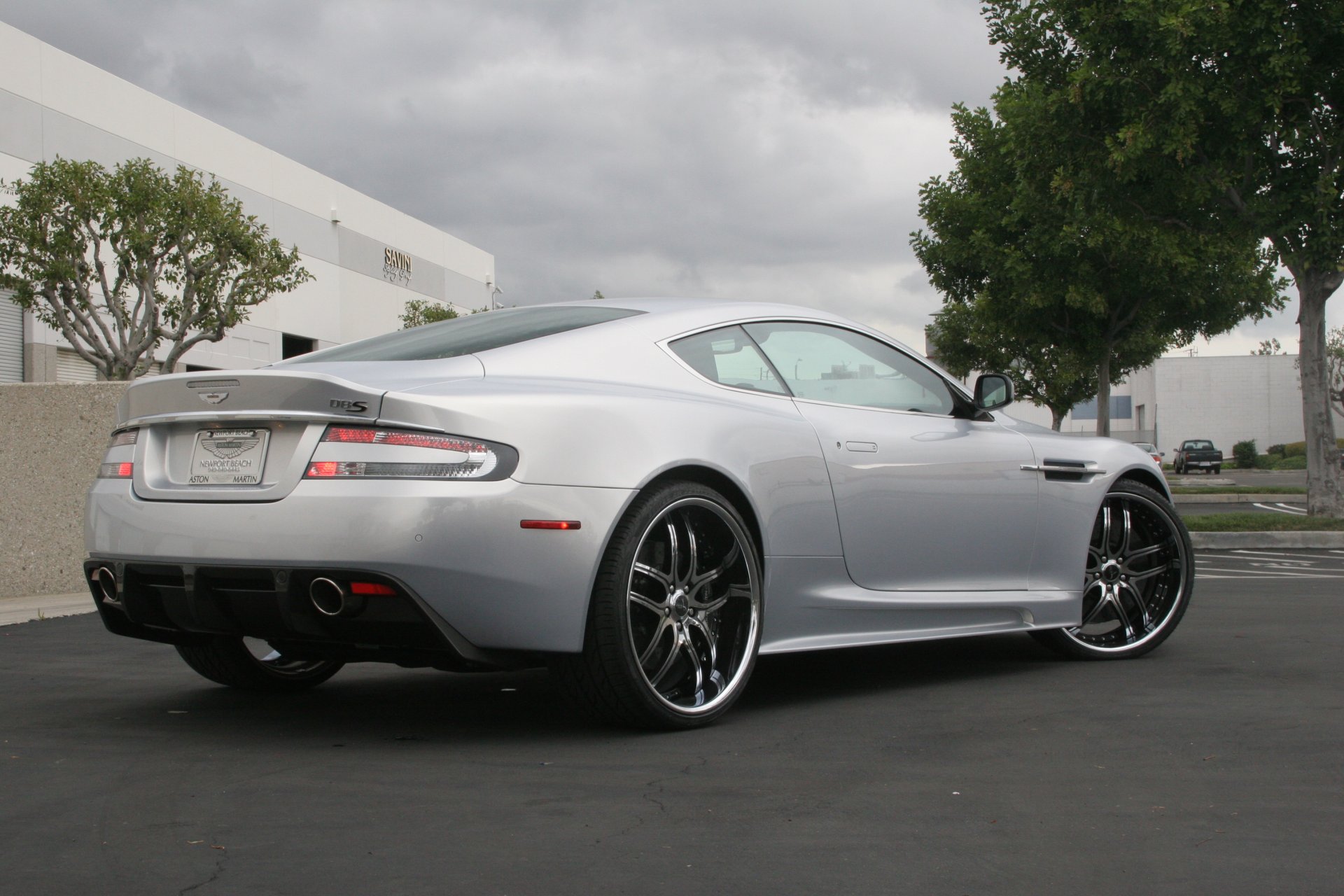 aston martin dbs argenté roues aston martin dbs argent vue arrière phare ciel nuages arbres bâtiment