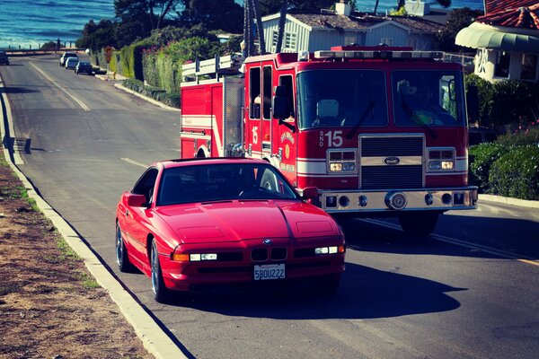 A red BMW car is driving next to a fire truck on the road