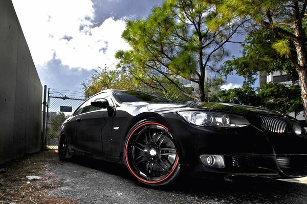 Photo of a black BMW on the background of a fence, pine trees and a blue sky with clouds