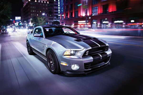 Grauer Ford Mustang mit zwei schwarzen Streifen auf der Motorhaube vor Nachtstadthintergrund