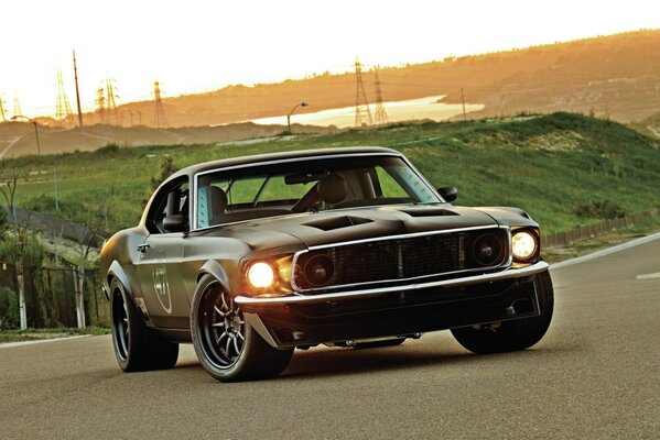 Ford Mustang noir avec phares allumés sur fond de coucher de soleil