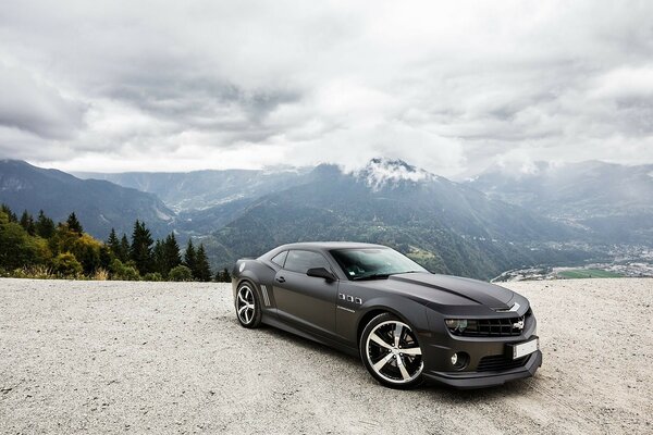 Black Camaro Chevrolet car on the road against the background of forest mountains and cloudy sky