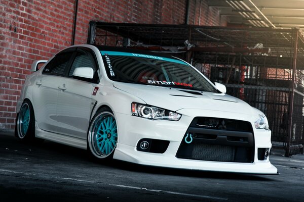 A white mitsubishi lancer car with blue tires stands against a brick wall