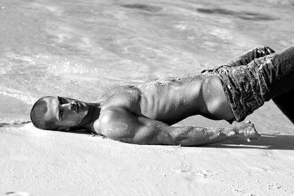 Schwarzweißes Foto eines schönen muskulösen Kerls in einer erotischen Pose im Sand