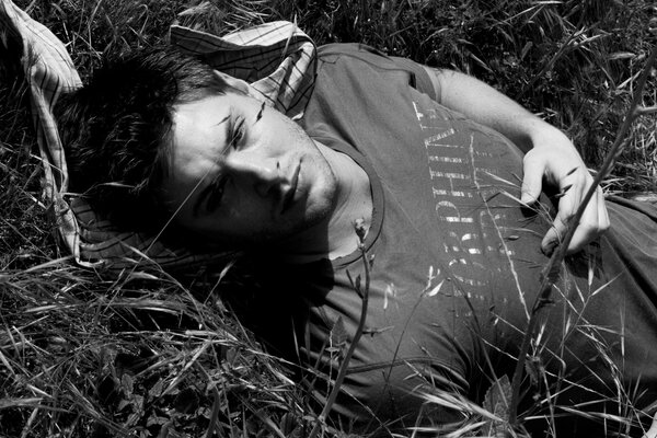 Ein nachdenklicher, dunkelhaariger Typ, der auf dem Rasen liegt. Dunkelhaarige hübscher Kerl auf einem Schwarz-Weiß-Foto, das im Gras liegt