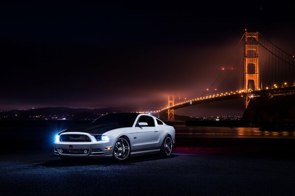En primer plano, en la noche, un Ford Mustang blanco cerca del puente iluminado con linternas vista frontal