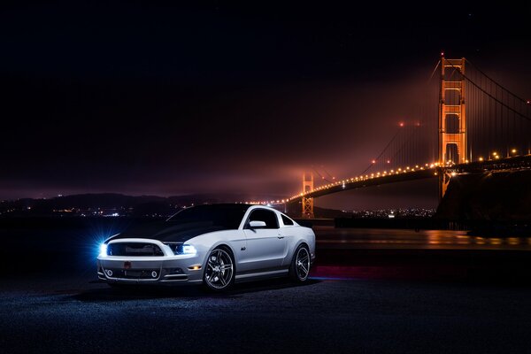 Au premier plan dans la nuit, une Ford Mustang blanche près du pont éclairé par des lanternes