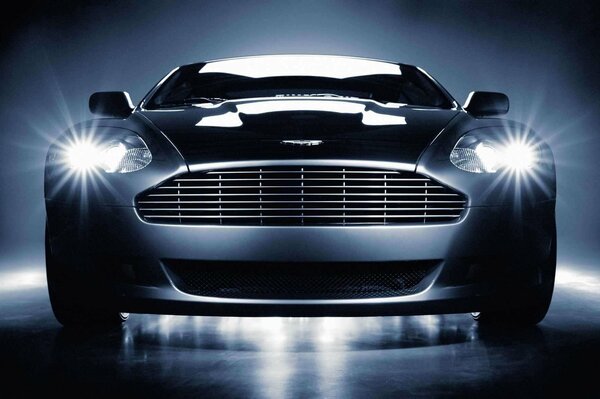 Czarno-biały obraz Aston Martin z włączonymi reflektorami. Aston Martin widok z przodu z włączonymi reflektorami