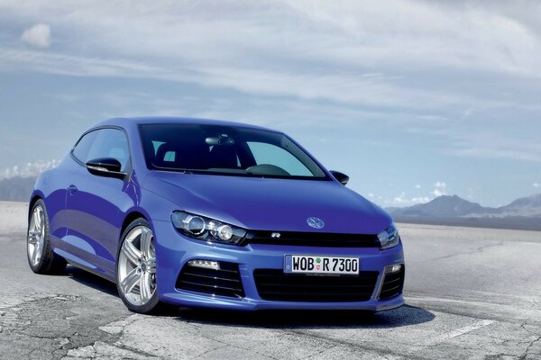 Volkswagen Scirocco blu su uno sfondo di montagne blu sotto un cielo nuvoloso