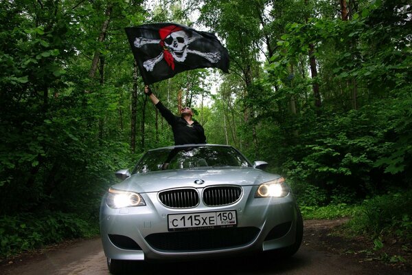 Hombre con bandera pirata en un BMW gris en un camino forestal