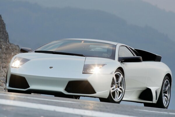 A white Lamborghini is driving on the road with its headlights on