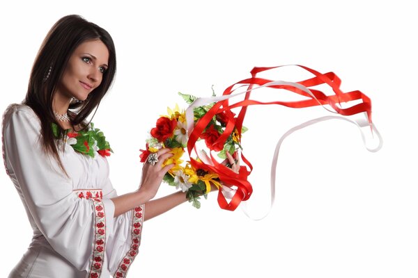 A girl in an embroidered white shirt holds a wreath with ribbons