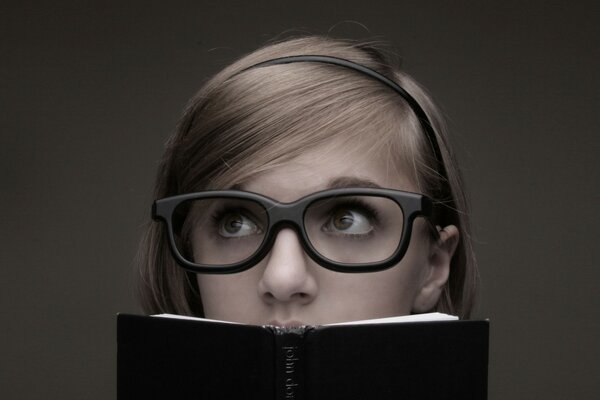 A girl with massive glasses looks over the top of a book