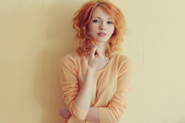 Red curls are sorted by a girl in a peach sweater against a cream wall