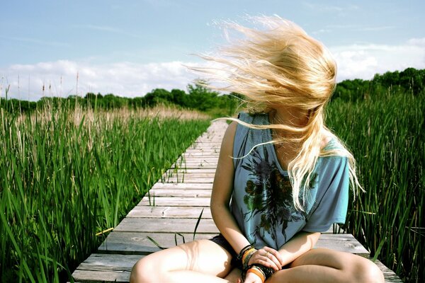 A blonde girl is sitting on a path among the grass and her hair is blown by a gust of wind