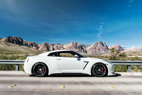 A white Nissan stands on the asphalt against the backdrop of mountains