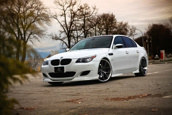 A tinted white bmw e60 sedan is parked on the asphalt in autumn