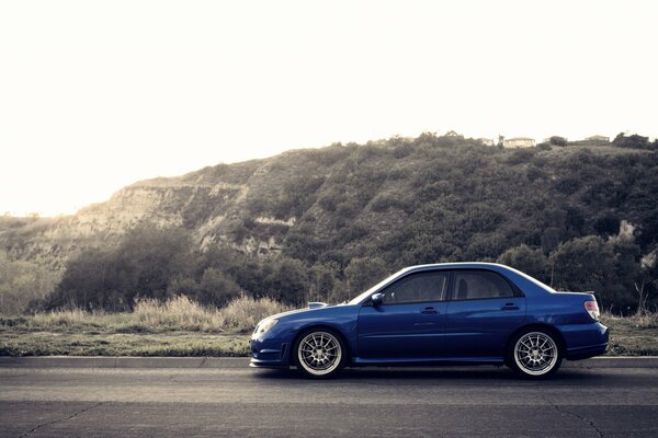 A blue Subaru car is driving on the road near the hills