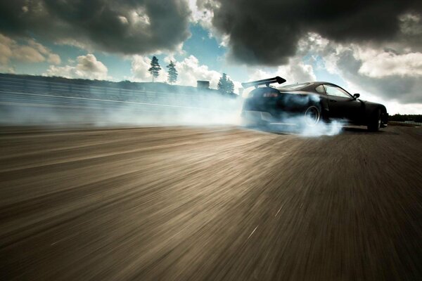 La deriva de un coche negro en el fondo de una pista de grafito y bajo un cielo nublado con nubes negras de humo