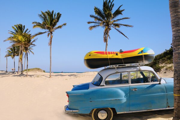 Voiture bleue soviétique avec un bateau sur le toit sur la plage de sable de l océan sous les palmiers sur fond de ciel bleu