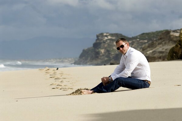 Daniel Craig mit dunkler Brille sitzt barfuß am Strand