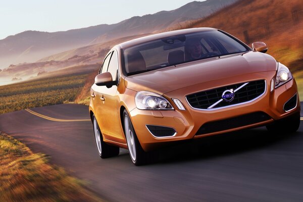 Auto Volvo arancione sulla strada nel campo contro uno sfondo di montagne e cielo blu chiaro