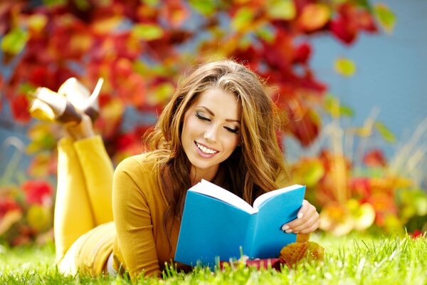 A young girl is reading a book lying on the green grass