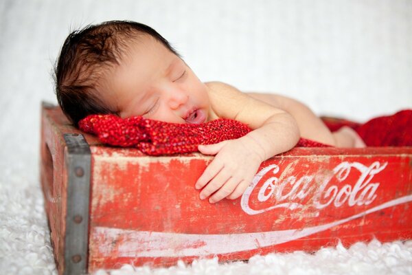 A sleeping child in a Coca-Cola box. Baby on a red blanket