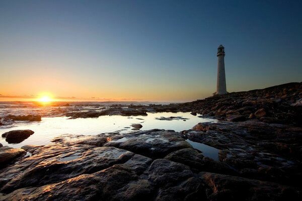 On the seashore there is a lighthouse on the background of a beautiful sun