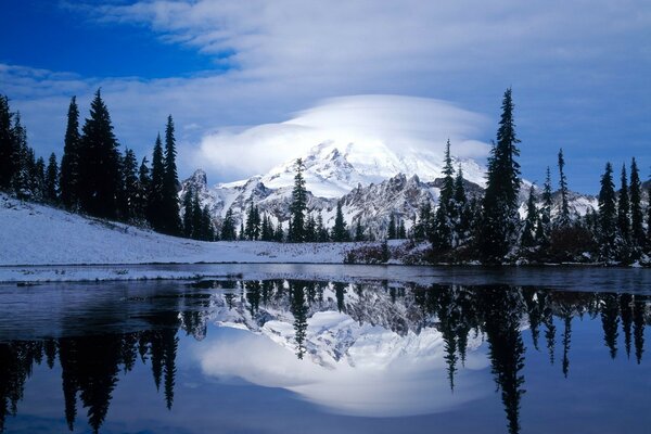 A snow-covered volcano in the clouds and trees are reflected in the lake