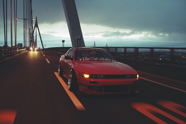 A red Nissan rides over a suspension bridge at sunset