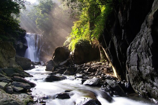 The sun s rays illuminate the waterfall among the stones