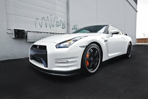 The front of a white Nissan parked on the asphalt against a gray wall with graffiti