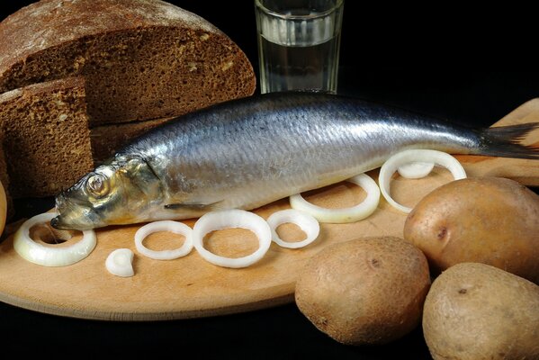 There are onion rings on the board, there are potatoes on the table next to it, there is black bread and there is a herring on the board