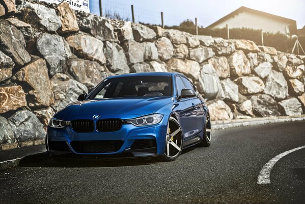 Un bmw azul tuneado en una curva contra un muro de piedra