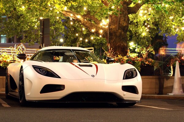 Un hipercoche blanco Koenigsegg Agera bajo un árbol con una guirnalda