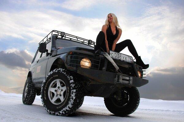 A beautiful blonde is sitting on the hood of a field