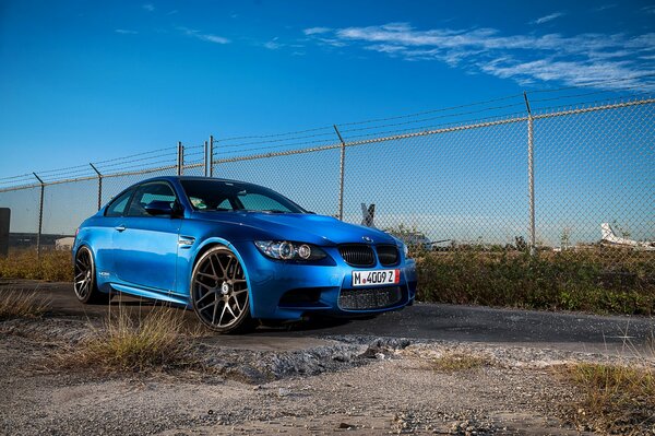 A beautiful BMW m3, blue as the sky, lurked near the fence