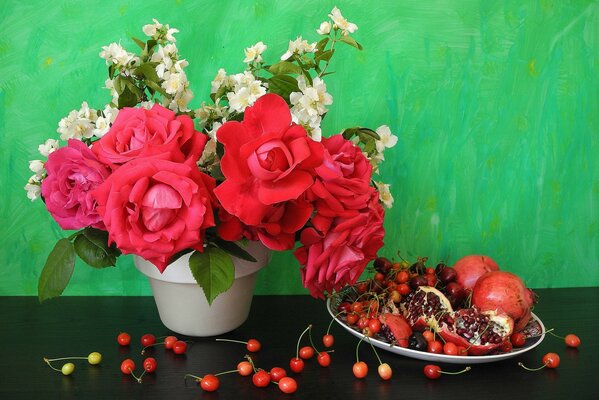 Sul tavolo c è una pentola di rose e un piatto di melograni e ciliegie