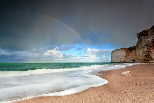 Das grüne Meer, die geschäumte Brandung und die sandige Küste mit einem Felsen vor dem Hintergrund eines nebligen Regenbogens