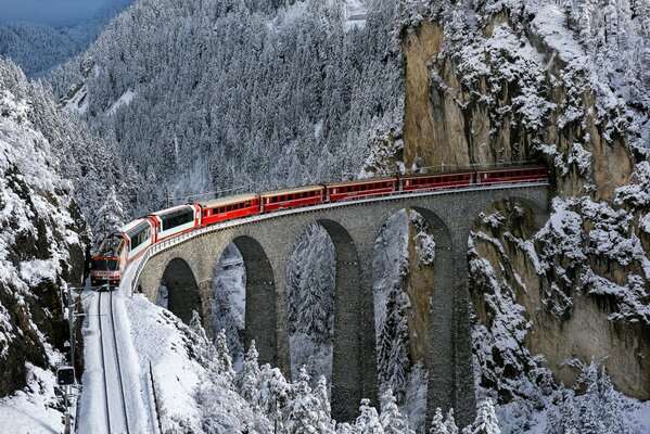 Ein roter Zug fährt aus einem Tunnel in einem Felsen auf einer schmalen, hohen Brücke, die inmitten eines schneebedeckten Bergwaldes steht