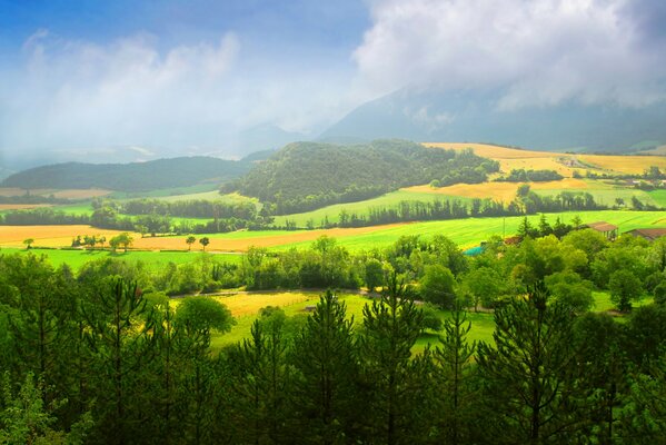 Zielone pola, domy i drzewa na tle gór wznoszących się szczytami w niebo