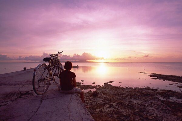 A girl with a bicycle on the background of a pink sunset in the reflection of water