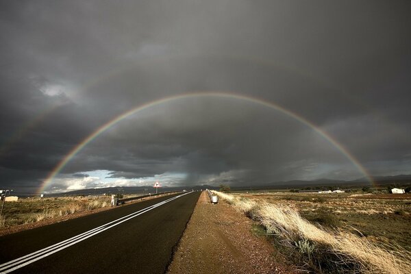 Die Straße, die unter dem Regenbogen vor dem Hintergrund von Wolken läuft