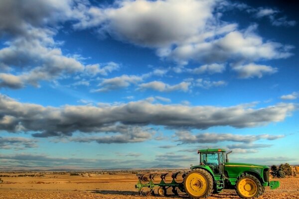 Ein grüner Traktor mit gelben Rädern steht inmitten eines Terrakottafeldes unter niedrigen Wolken am blauen Himmel