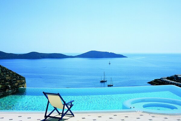 A chair by the azure pool located next to the sea where yachts sail and mountains are visible and the sea itself merges with the sky