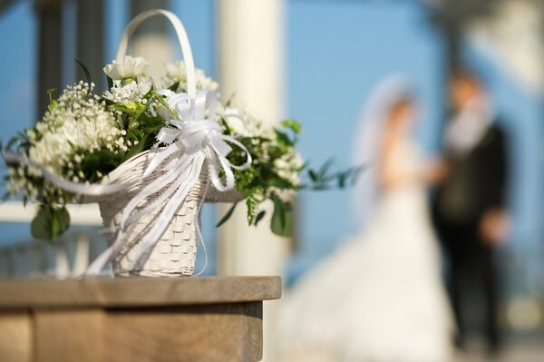 Cesta de flores blancas de la boda con la cinta blanca en el fondo de la novia y el novio borrosos